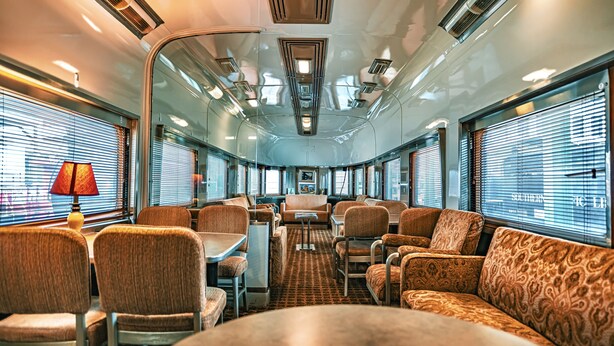 Interior de un tren con asientos, mesas y una mesa central con sillas.