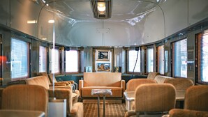 Interior de un tren con una zona de asientos centrales, mesas y ventanas amplias.