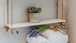 A white shelf with plants and jars, tied with rope, and a stack of towels with lavender. - Bay Tree Cottage - Luxury Cottages (Cheltenham)