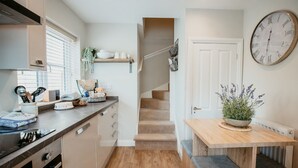 A modern kitchen with a built-in oven, a dining area with a wooden table and bench, and a staircase leading to another level. - Bay Tree Cottage - Luxury Cottages (Cheltenham)