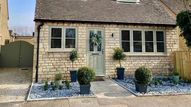 A two-story stone house with a gabled roof, a front door with a wreath, and a small garden with potted plants. - Bay Tree Cottage - Luxury Cottages (Cheltenham)