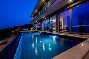 A modern poolside area with a clear view of the pool, a seating area with lounge chairs, and a large glass door leading to the interior. - Villa Semper Fi with Pool (Opatija)