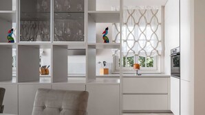 A modern kitchen with white cabinetry, a dining area with a grey upholstered chair, and a window with a geometric curtain. - Villa Palazzo di Cleo with Pool (Vabriga)