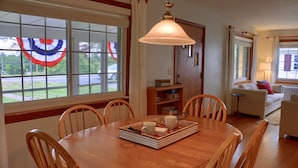 A dining area with a wooden table, chairs, and a tray with cups and a book. There is a window with curtains and a view of the outside. - Visit our charming cottage this Fall & Winter. Enjoy the Scenery & Ski Slopes. (McKnightstown)