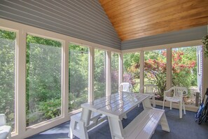 Relax in this serene sunroom with a view of lush greenery and flowering plants, featuring a wooden table and comfortable seating.