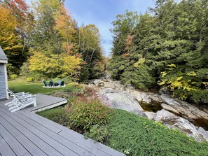 A deck with outdoor seating, a small stream, and surrounding trees with autumn foliage.