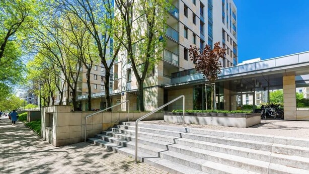 Un edificio moderno con una escalera que conduce a un acceso cubierto, rodeado de árboles y un cielo despejado.