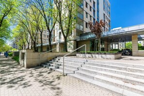 A modern building with a staircase leading to a covered entrance, surrounded by trees and a clear sky.