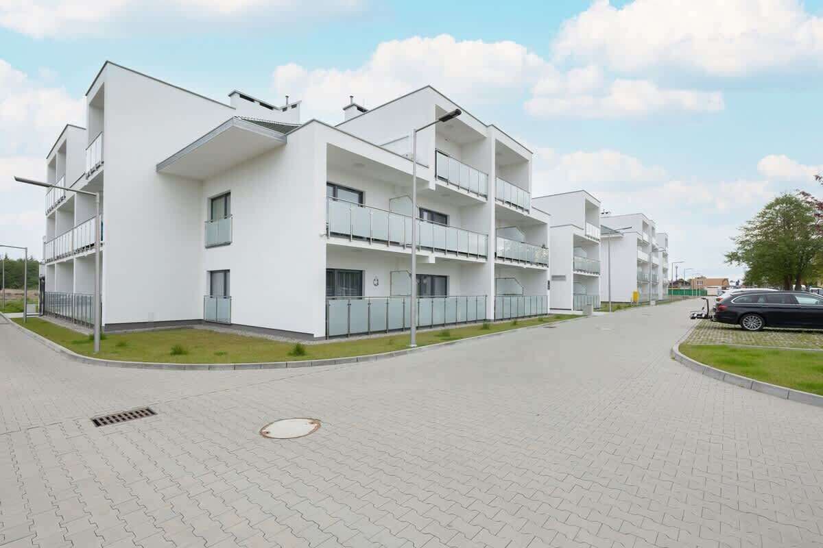 Um edifício residencial moderno de vários andares, com varandas, entrada pavimentada e um carro estacionado perto.