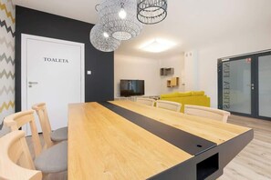 En moderne stue med et langt træbord, to sofaer, et fjernsyn og en dør med teksten 'TOALETA'.