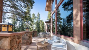Un espace patio avec des murs en pierre, une table ronde, des chaises blanches et une vue sur de grands arbres, visible à travers de larges fenêtres.