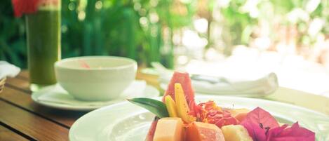 A table with a plate of fruit and a glass of green drink, set outdoors with greenery in the background.