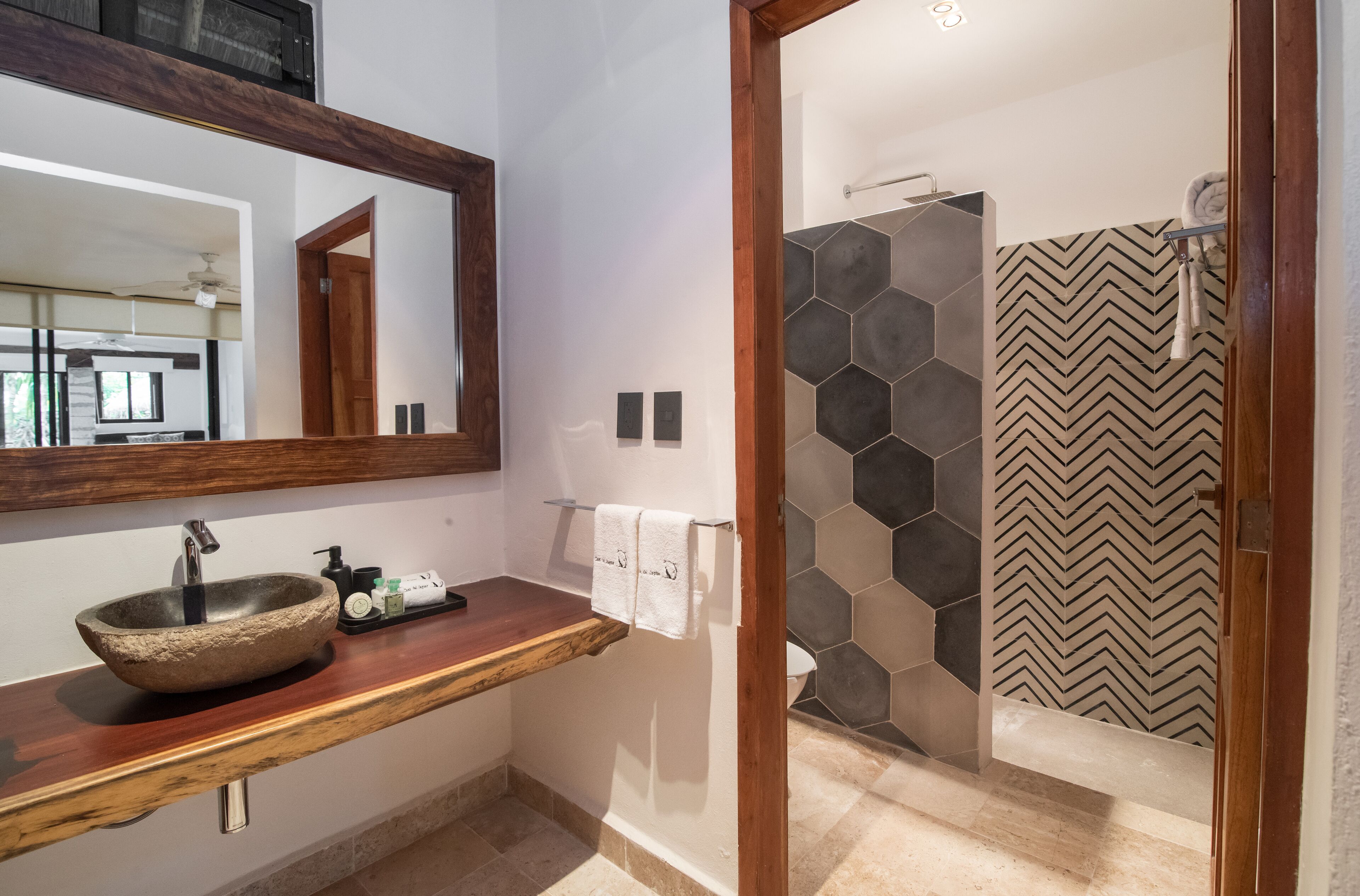 Un baño moderno con tocador de madera, un espejo grande, lavabo sobre encimera y zona de ducha con azulejos estampados.