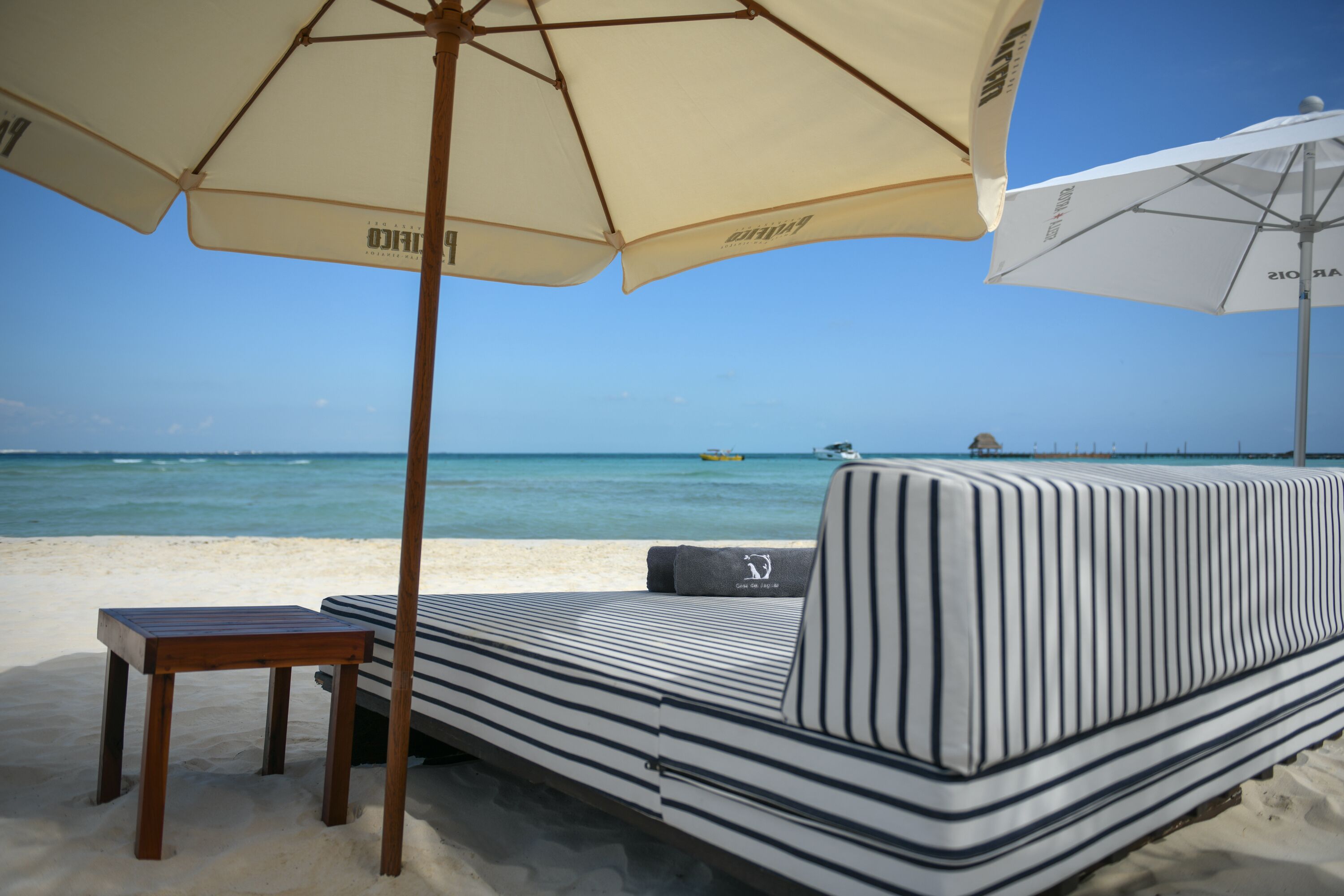 A beachside lounge with a striped sofa and a small wooden table, an umbrella providing shade, and a view of the ocean.