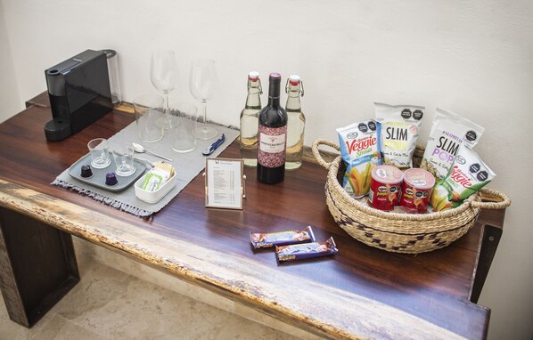 Una mesa de madera con una cafetera, copas de vino, botellas, aperitivos y una cesta.