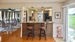 A modern kitchen with a bar area, wooden flooring, and a dining area with a glass table and chairs. - Burnt Store Isles Golf Home, W/Hot Tub/King Bed/Fast WIFI/Unbeatable Location (Punta Gorda)