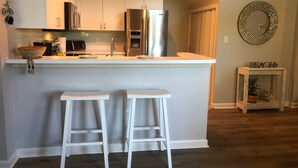 Una cocina con armarios blancos, un refrigerador de acero inoxidable, un microondas y un lavamanos. Hay dos taburetes blancos frente a la encimera.