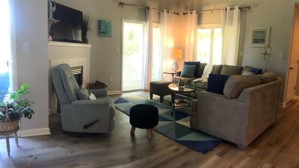 Una sala de estar con un sillón gris, un sofá, una mesita de centro y un tapete estampado. Hay un televisor montado en la pared y una ventana con cortinas.