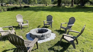 Un espace extérieur verdoyant, aménagé avec des fauteuils Adirondack disposés autour d’un foyer en pierre.