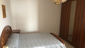 Una cama bien hecha con una colcha estampada, una mesita de noche con una lámpara y un armario de madera en una habitación con suelo de baldosas.