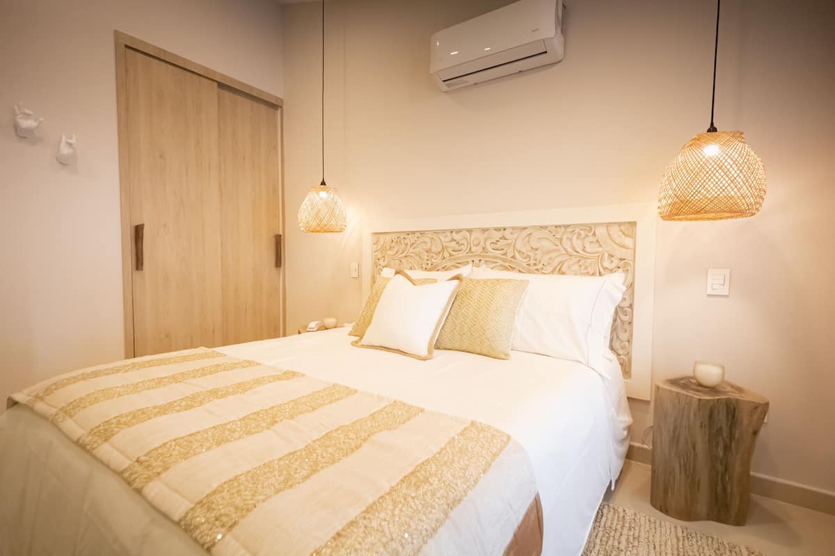Een keurig opgemaakt bed met een gestreept dekbedovertrek, een houten hoofdbord met een decoratief patroon, en twee hanglampen met een geweven ontwerp.