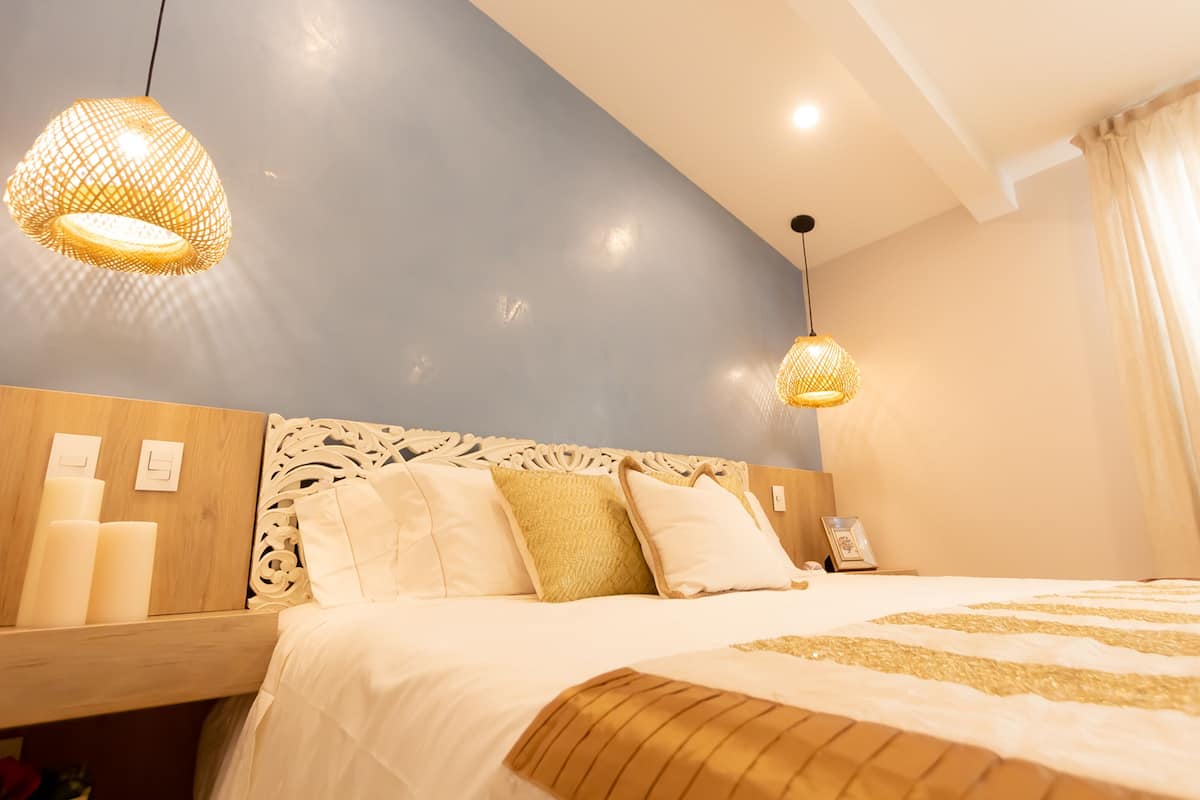 Een net opgemaakt bed met wit en goud linnengoed, een houten hoofdbord met ingewikkelde gravures, en twee hanglampen met een geweven ontwerp.