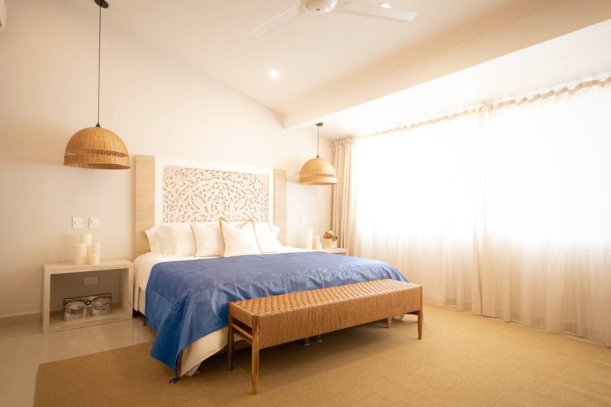 Een slaapkamer met een bed, nachtkastjes, een bankje en een plafondventilator.