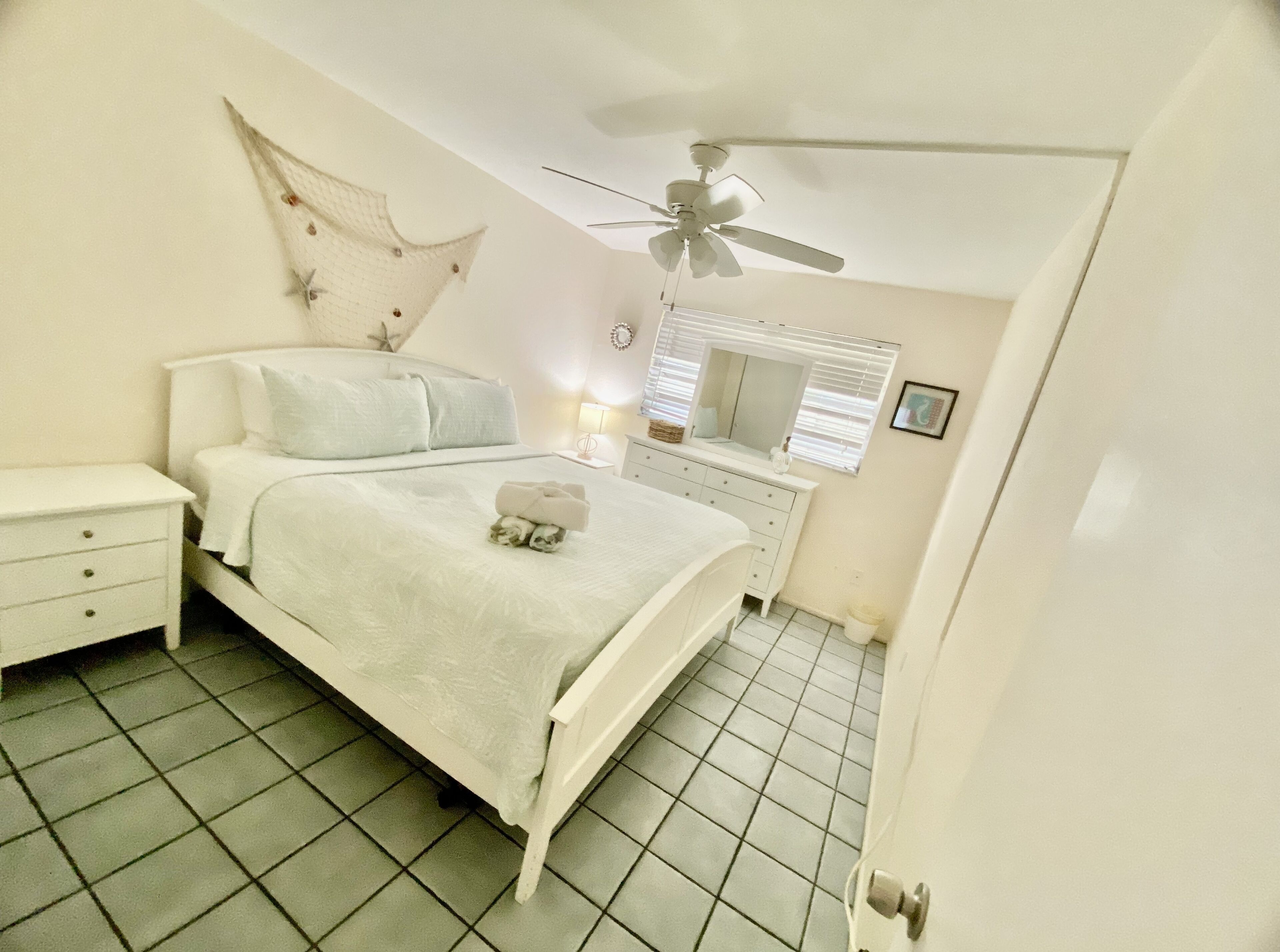 Une chambre à coucher comprenant un lit, une table de chevet, une commode, un miroir, un ventilateur de plafond et un sol carrelé.