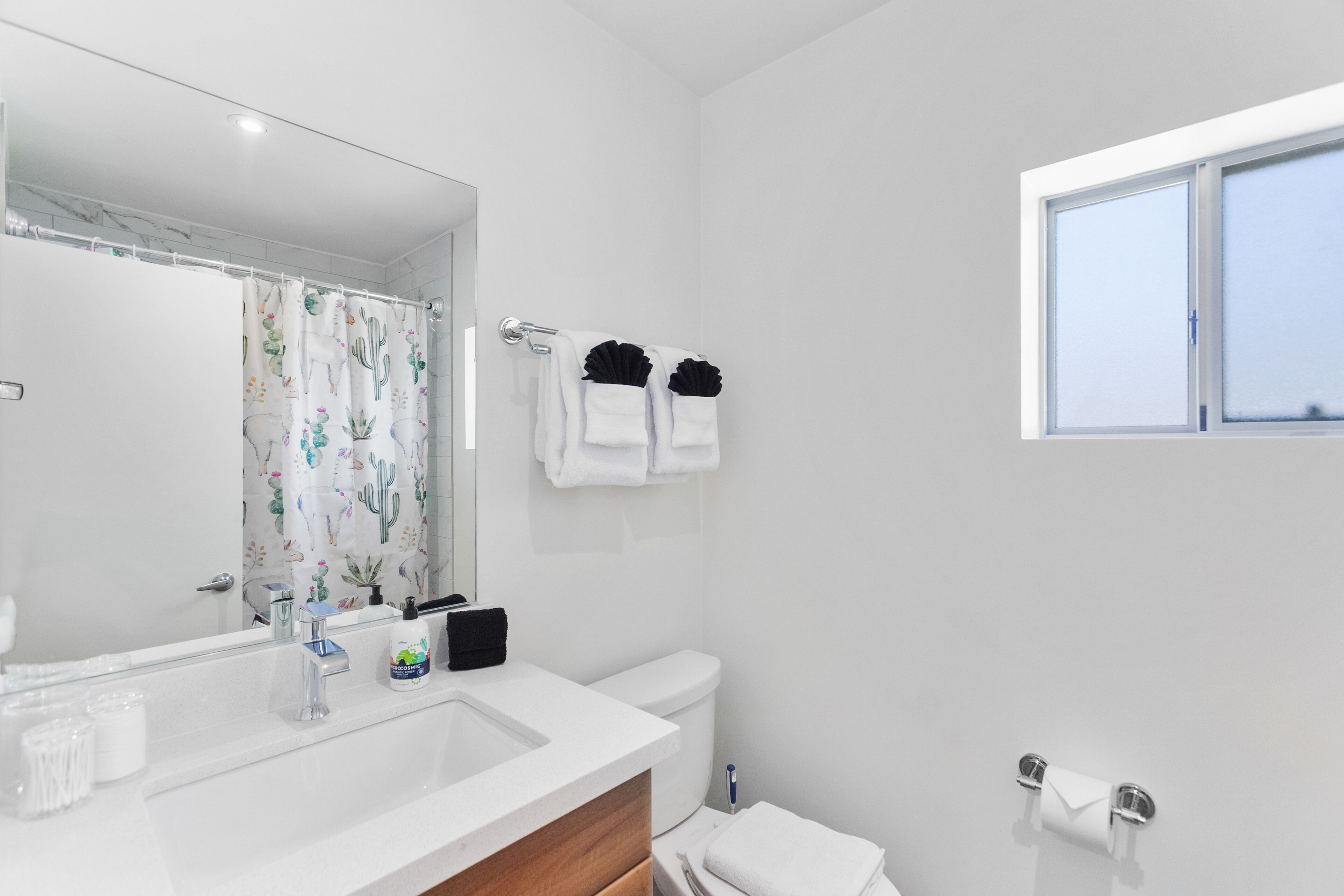 A modern bathroom with a white sink, mirror, and toilet. There is a window providing natural light and a shower curtain with a cactus design.