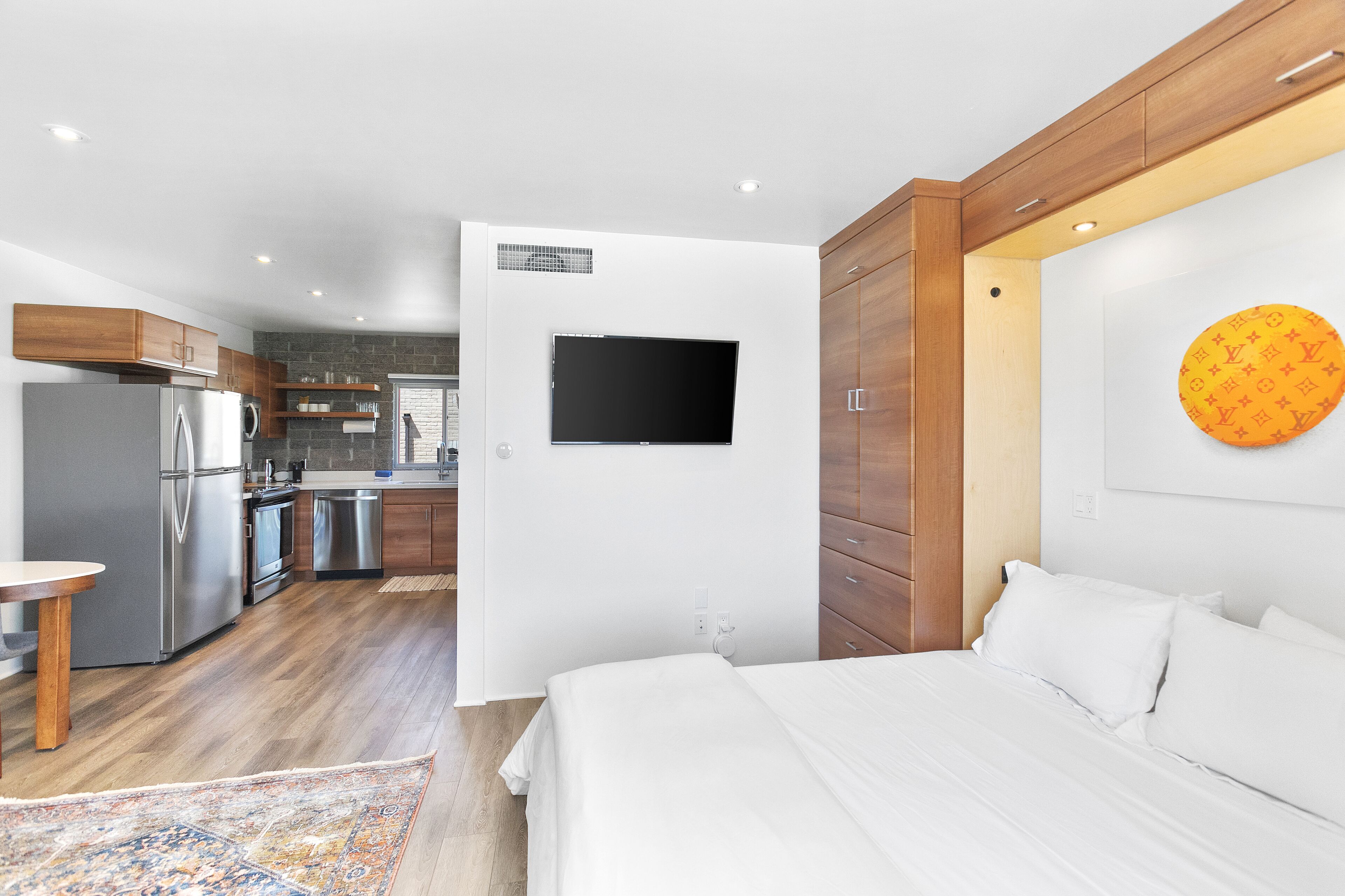 A modern hotel room with a large bed, a flat-screen TV mounted on the wall, a kitchen area with modern appliances, and a wooden ceiling.