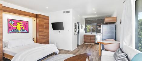 Une chambre moderne avec un lit, une petite table en bois et un canapé. On aperçoit un coin cuisine à l’arrière-plan.