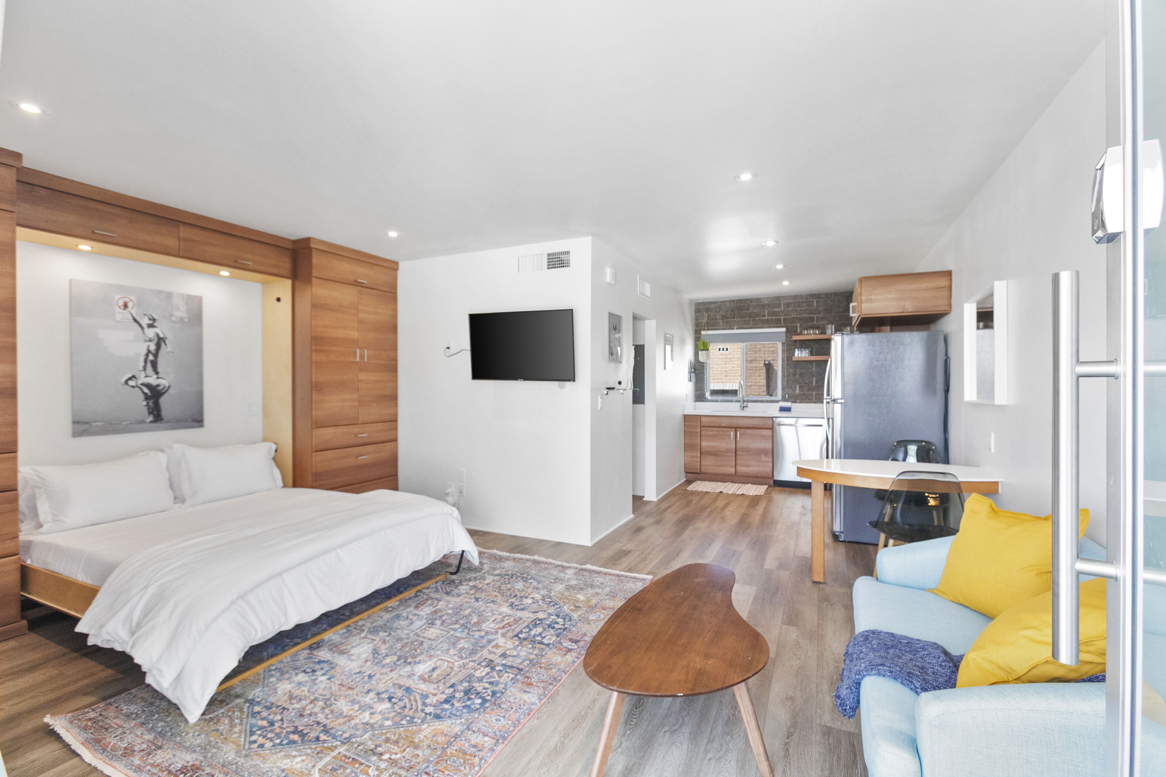 A modern bedroom with a large bed, a sitting area with a sofa, and a kitchen in the background.