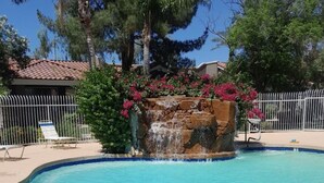 A swimming pool with a waterfall feature surrounded by lush greenery and a white fence.