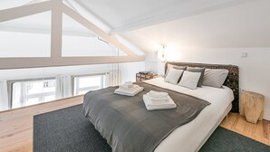 Een moderne slaapkamer met een schuine kap, een groot bed met een grijs en wit dekbed, meerdere kussens, een nachtkastje met boeken, en uitzicht op de stad door een groot raam.