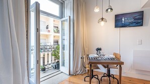 Een kamer met een houten eettafel gedekt voor twee personen, een aan de muur bevestigde televisie, en een balkon met een hekwerk en bloempotten.
