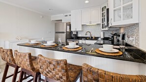 A modern kitchen with white cabinetry, a black countertop, and woven chairs. - Direct Oceanfront View- 213 Breakers-Pool (Hilton Head Island)