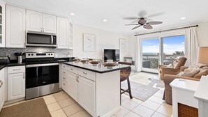 A modern kitchen with white cabinets, a built-in microwave, and a large island. There is a ceiling fan, a large window with a view, and a seating area with a sofa and a chair. - Direct Oceanfront View- 213 Breakers-Pool (Hilton Head Island)