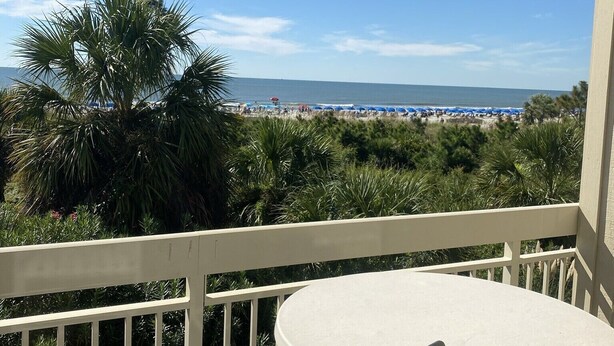 A balcony with a white table and chair, overlooking a beach with umbrellas and palm trees. - Direct Oceanfront View- 213 Breakers-Pool (Hilton Head Island)
