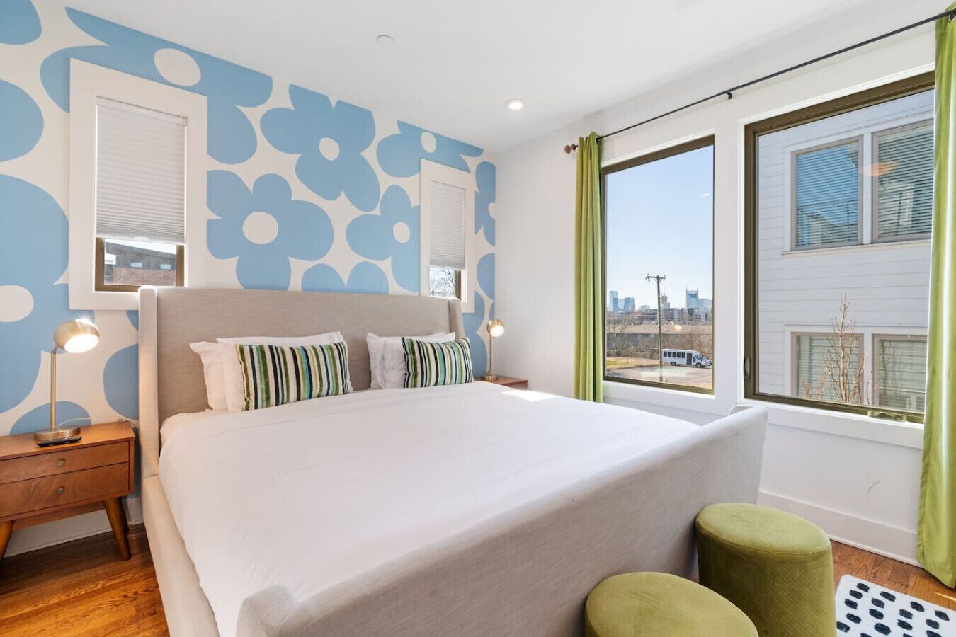 A modern bedroom with a large bed, a nightstand, a lamp, and a view of the city through a window.
