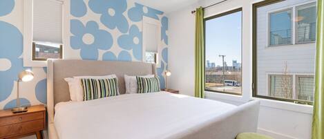 A modern bedroom with a large bed, a nightstand, a lamp, and a view of the city through a window.