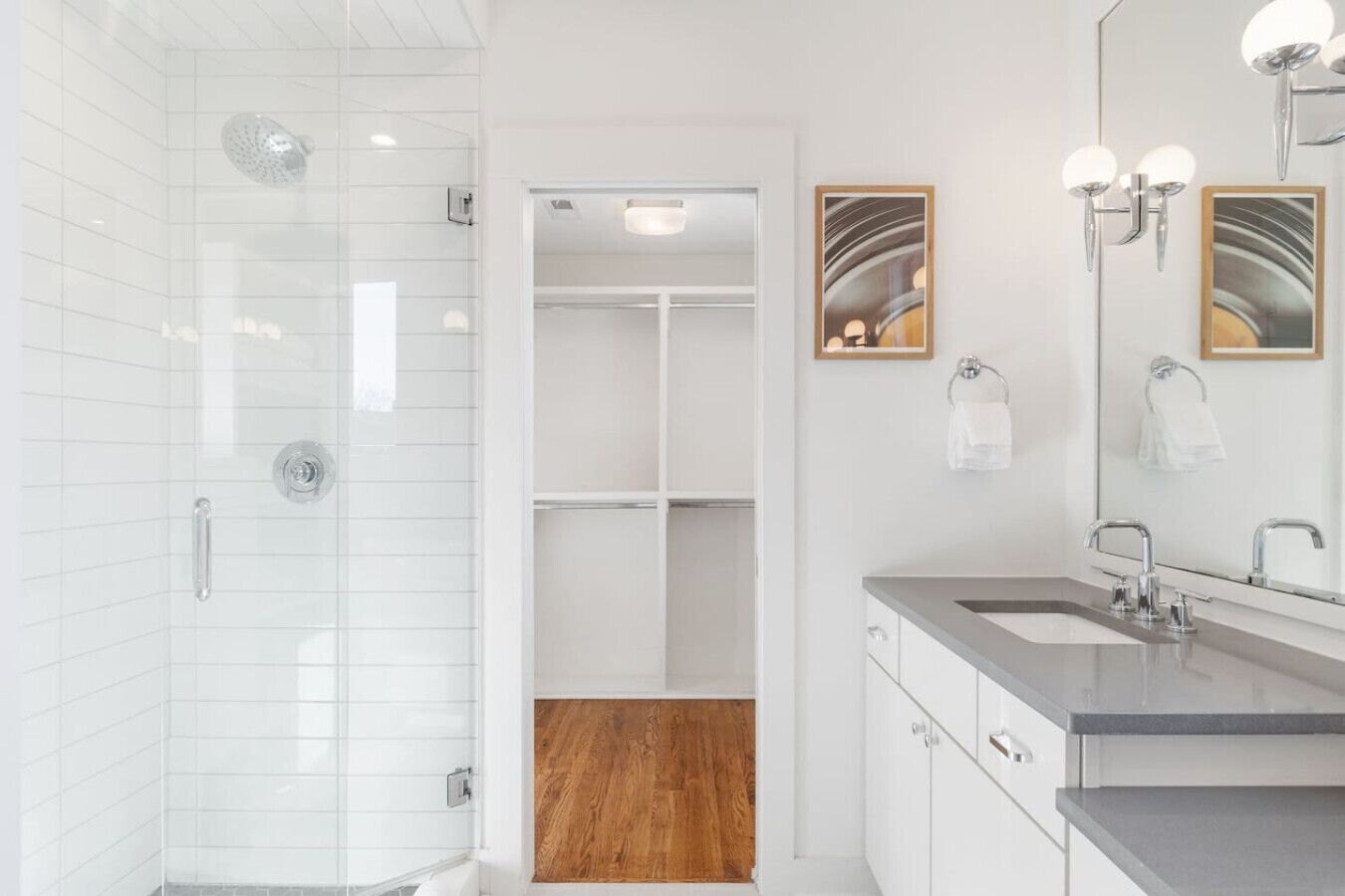 A modern bathroom with a walk-in shower, a vanity with a sink and mirror, and a wooden floor leading to a closet.
