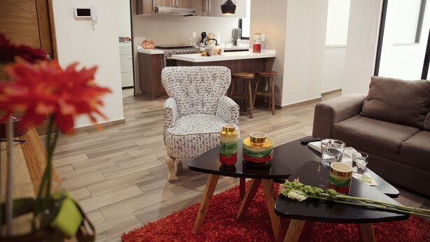 A modern living room with a sofa, a coffee table with decorative items, a kitchen area with wooden cabinets, and a red rug.