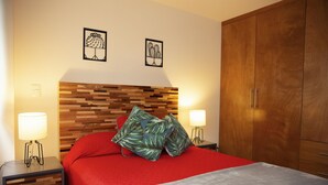A bedroom with a wooden headboard, red bedding, two wall-mounted lamps, and a wardrobe.
