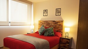 A neatly made bed with a red bedspread and pillows, a wooden headboard, bedside lamps, and two wall art pieces.