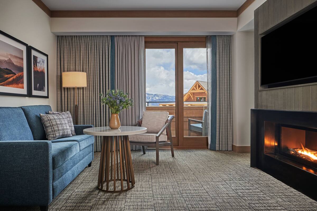 Ein Hotelzimmer mit blauem Sofa, einem runden Tisch mit Vase, einem Flachbildfernseher, einem Kamin und einem Balkon mit Bergblick.