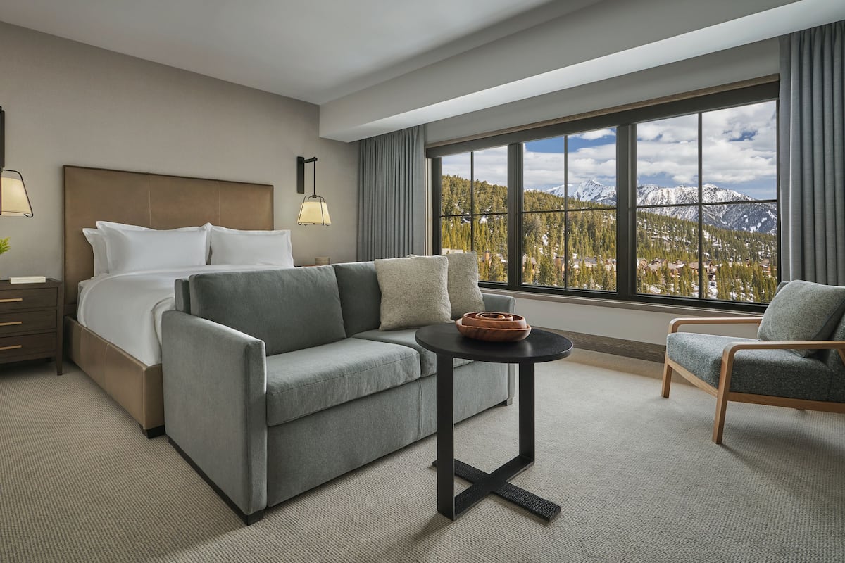Ein Hotelzimmer mit Bett, einer Couch, einem kleinen Tisch und einem Stuhl. Ein großes Fenster bietet Blick auf Berge und Bäume.