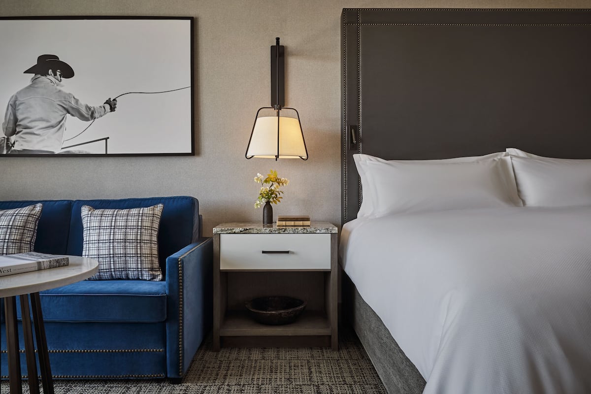 Ein Hotelzimmer mit einem Bett, einem blauen Sofa mit karierten Kissen, einem kleinen Tisch mit einem Buch, einem Nachttisch mit einer Lampe und einem gerahmten Bild an der Wand.