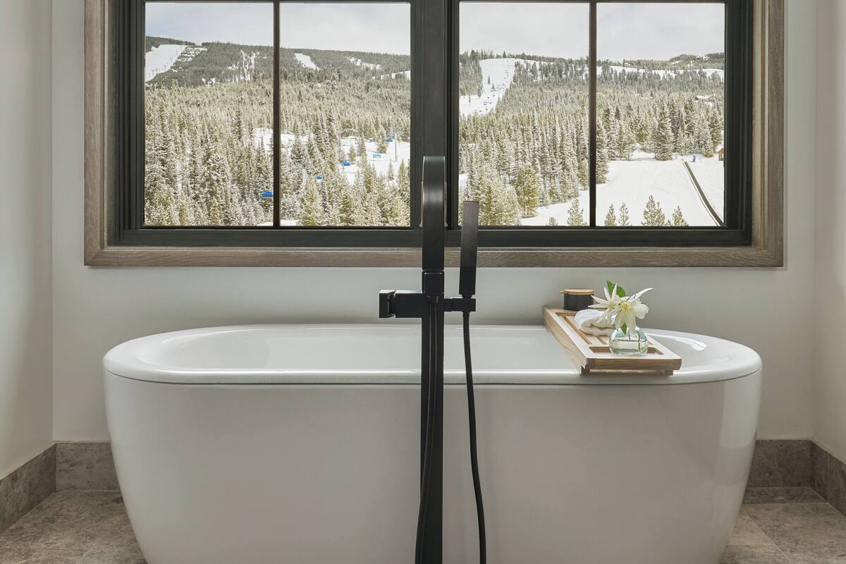 Ein Badezimmer mit einer freistehenden Badewanne, einem Fenster mit Blick auf eine verschneite Landschaft und einer kleinen Holztablett mit einer Blume auf der Badewanne.