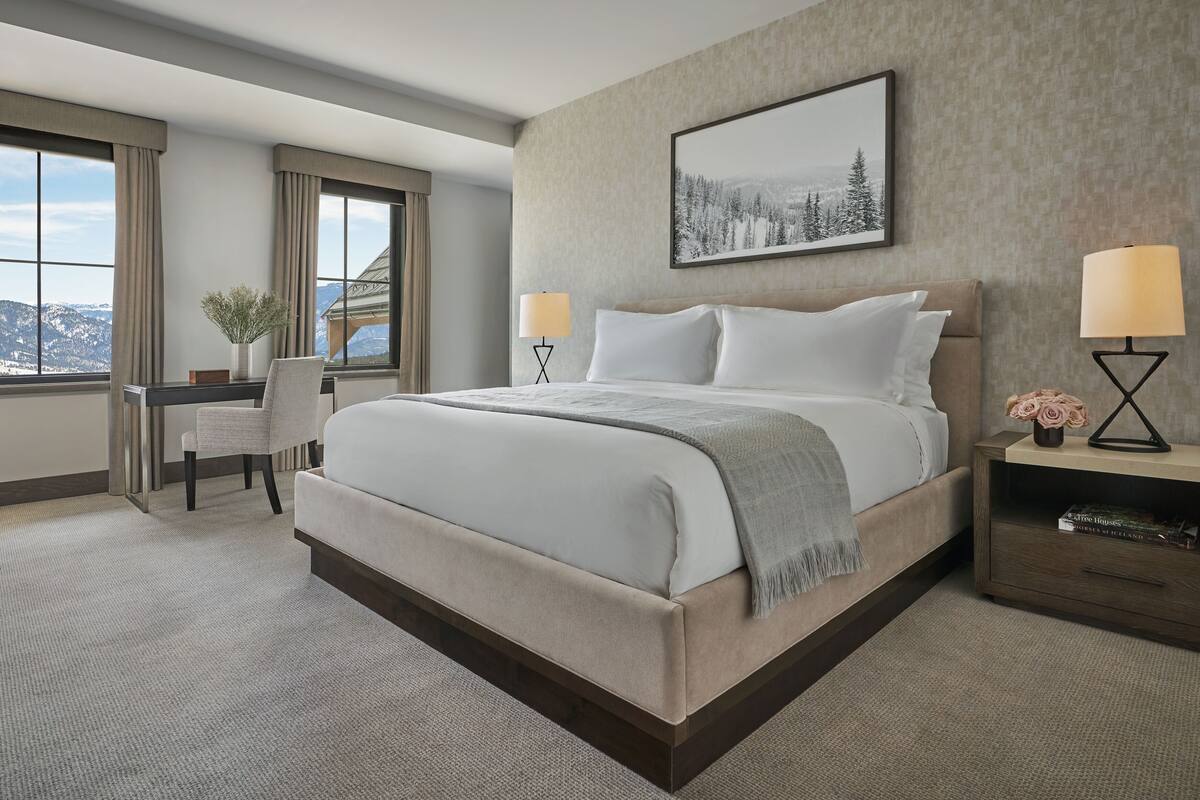 Ein Hotelzimmer mit einem großen Bett, zwei Nachttischlampen, einem Schreibtisch mit Stuhl, Blick auf Berge durch große Fenster und einem gerahmten Bild an der Wand.