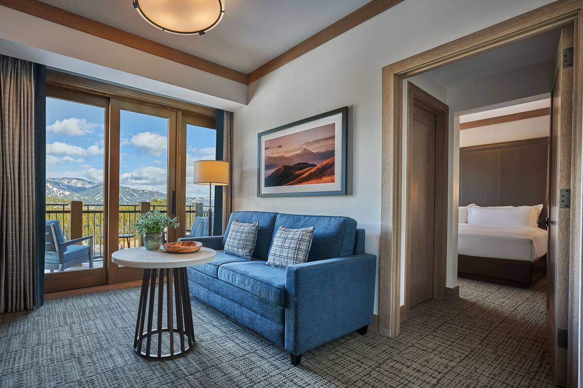 Ein Hotelzimmer mit einem blauen Sofa, einem kleinen runden Tisch mit Schüssel und Pflanze, einem großen Fenster mit Bergblick, einem gerahmten Bild an der Wand und einem Bett mit weißer Bettwäsche.